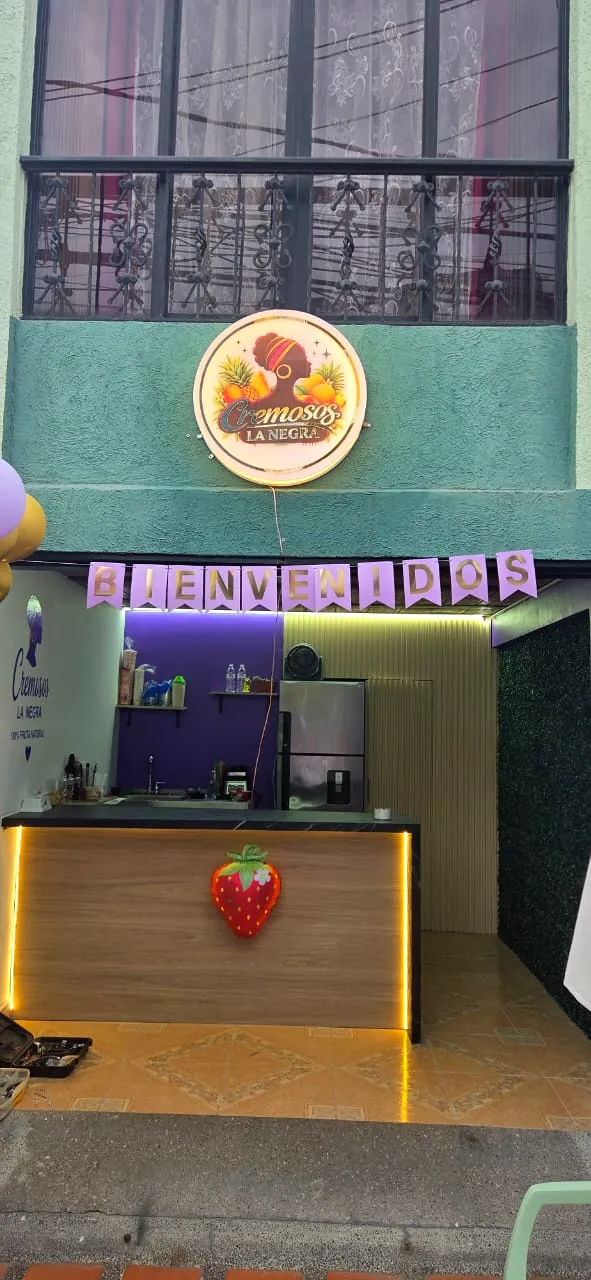 Fachada verde con el aviso redondo iluminado de Cremosos La Negra y un banderín de Bienvenidos