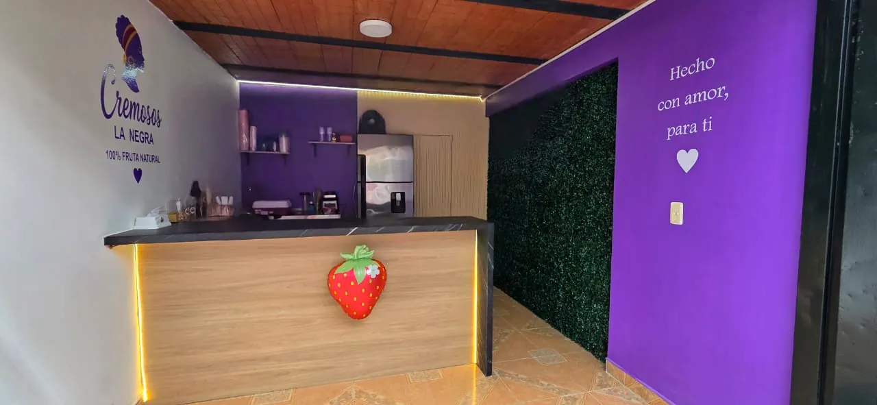 Interior de Cremosos La Negra: barra de madera con luz dorada, una fresa gigante, pared morada con la frase Hecho con amor, para ti y muro verde